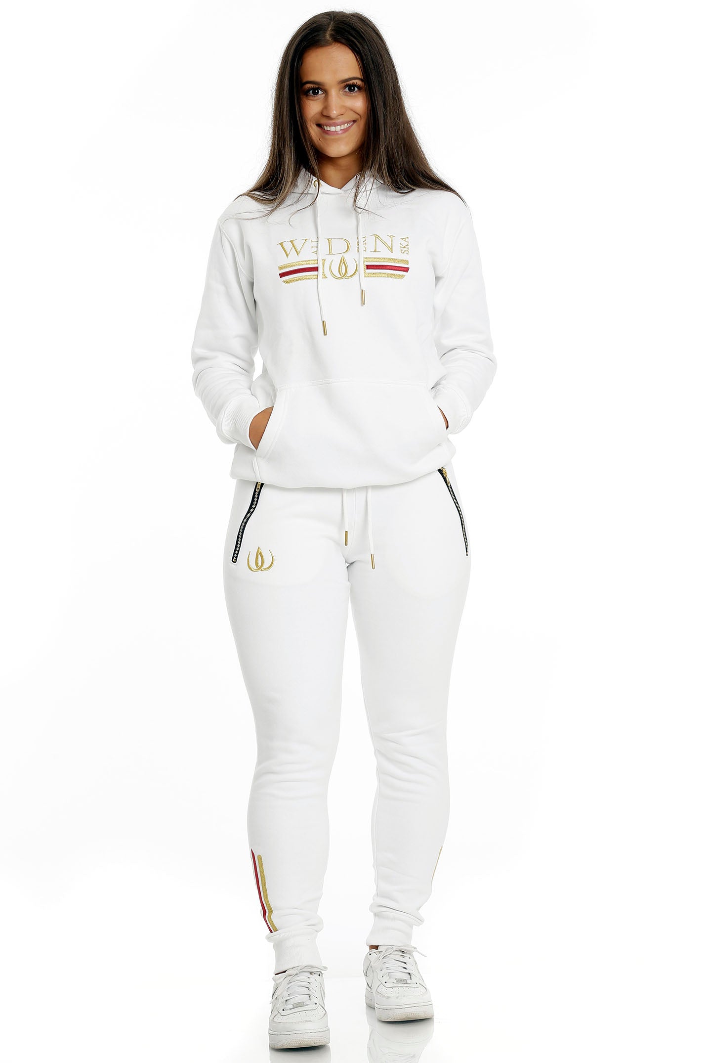 Staand in witte joggers met gouden logo en witte hoodie met goudborduursel, gedragen met witte sneakers.