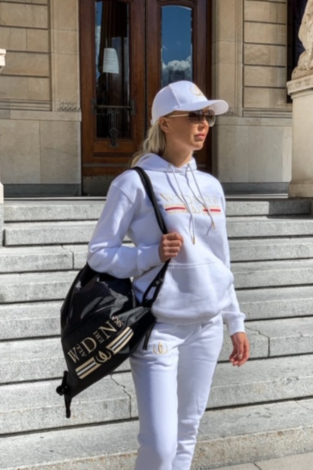 De joggers worden gedragen met een witte hoodie en pet, met een zwarte rugzak met gouden logo over de schouder.
