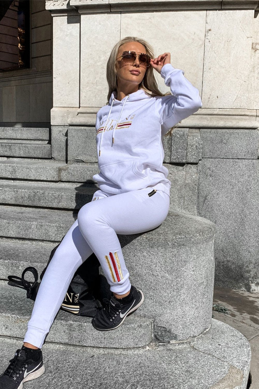 Op een stenen trap in de stad draagt het model witte joggers met goudstreep bij de enkel en een witte hoodie.