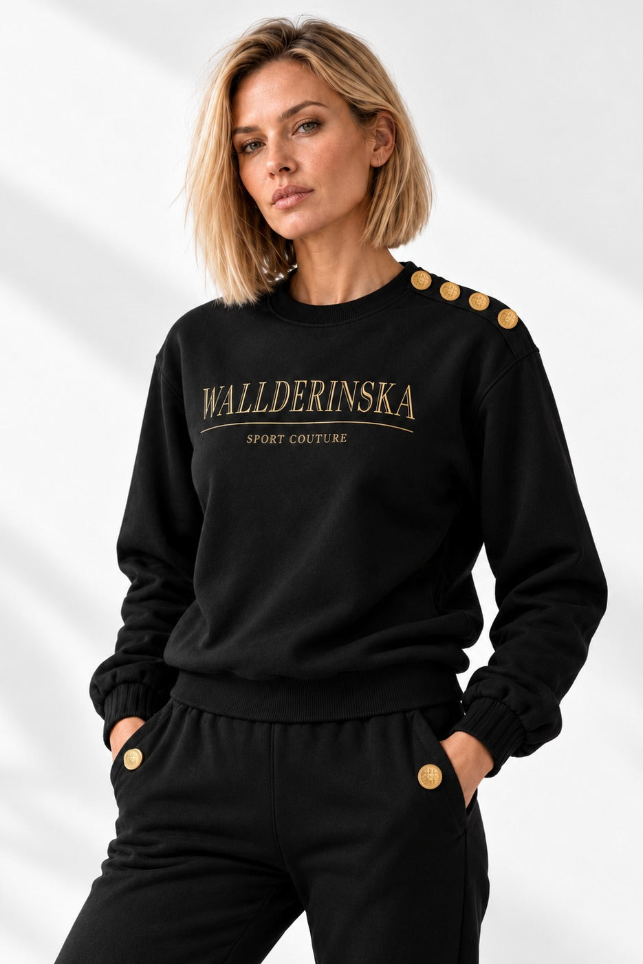 Edge Gold Button Sweater i svart framifrån i ljus studio, med fyra guldknappar på axeln och guldtext på bröstet.