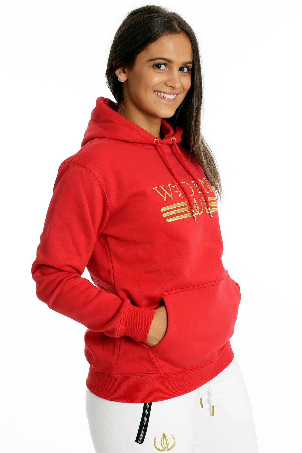 De handen rusten in de buidelzak van de rode hoodie. Daaronder zijn witte joggers met goudkleurige rits te zien.