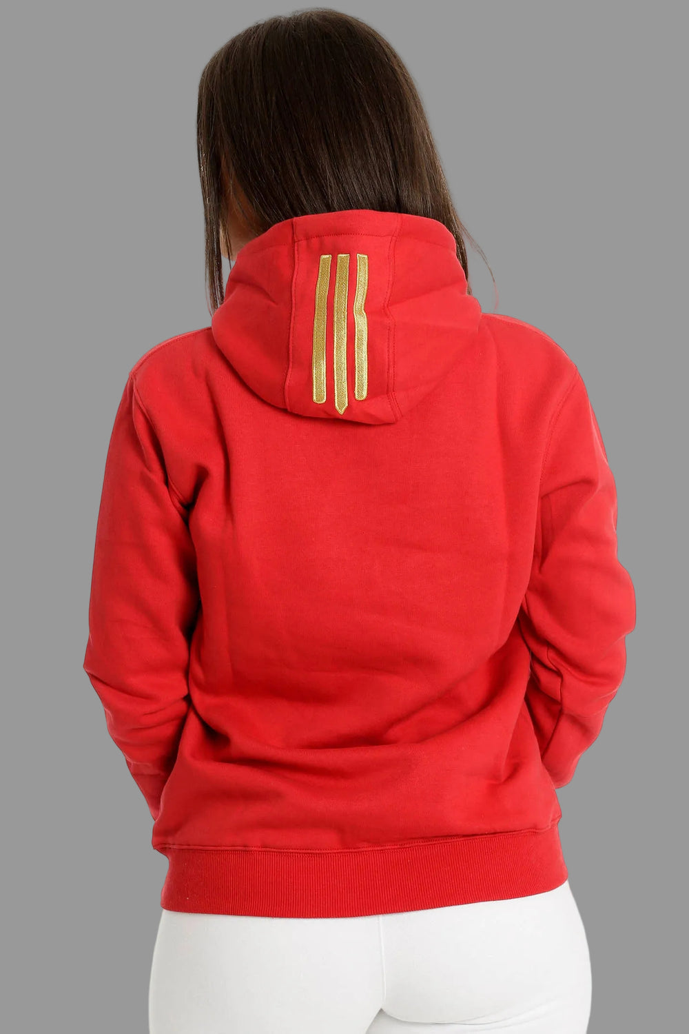 Achterkant van de rode hoodie met drie goudkleurige strepen op de capuchon. Het model draagt een witte broek.