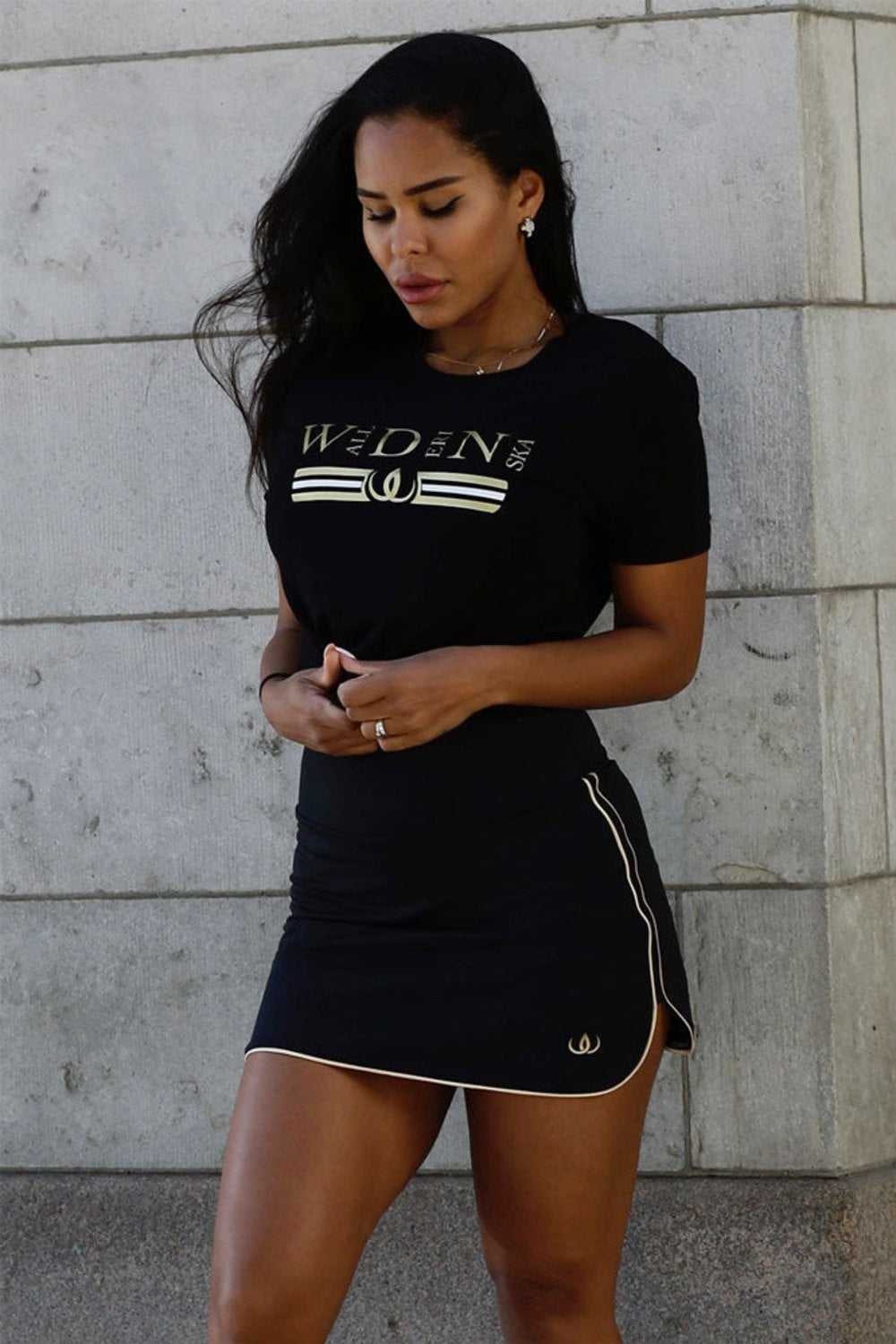 Het model staat tegen een lichte muur in de zwarte rok en een zwart t-shirt met gouden print.