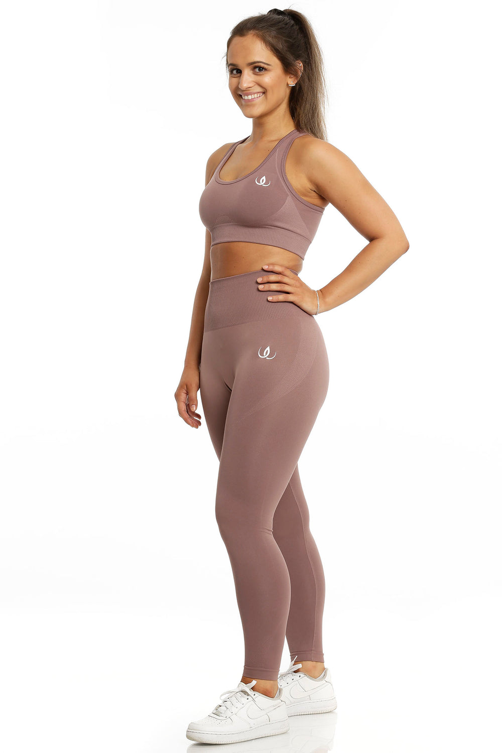 Sport-bh en legging in dezelfde rosébruine tint. Het model staat met de hand in de zij, in witte schoenen.