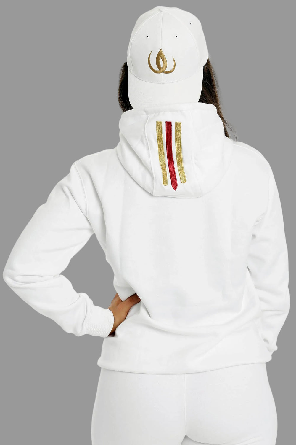 Achterkant van de witte hoodie met twee goudstrepen en een rode op de capuchon. Het model draagt een witte pet.