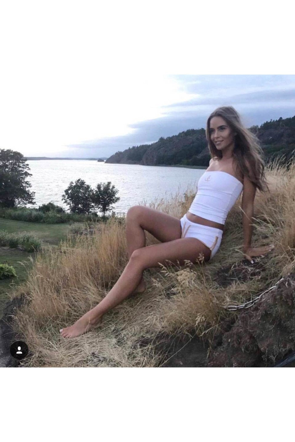 Het model leunt achterover in het gras bij het water, in een witte short met gouden rand en witte bandeautop.