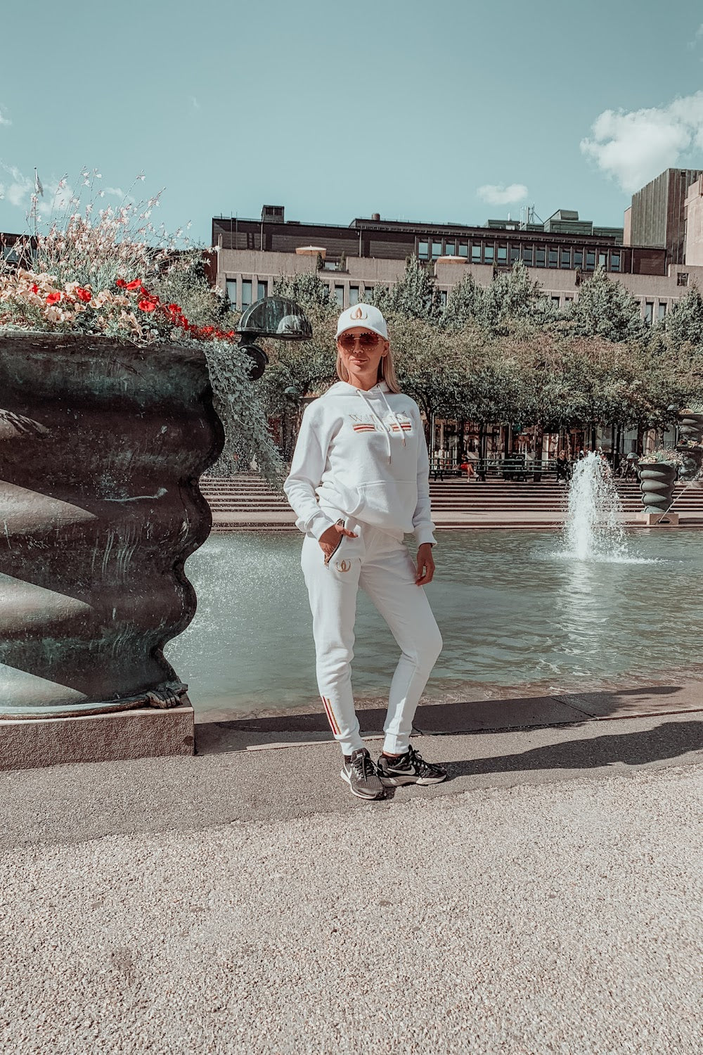 Voor de fontein staat het model in een witte hoodie, witte joggers en een witte pet.
