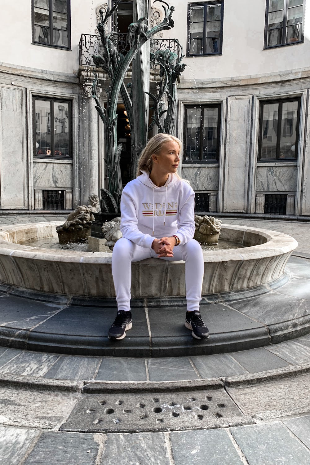 Op de rand van de fontein zit het model in de witte hoodie, met een standbeeld op de achtergrond.
