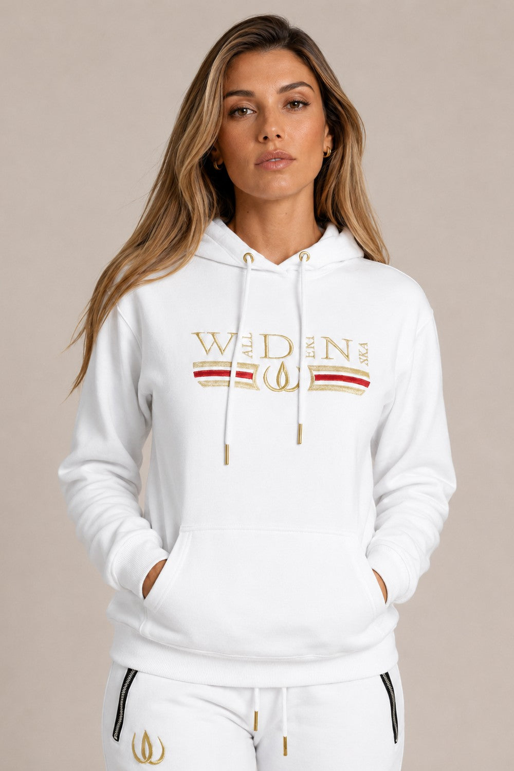 Witte Deluxe Hoodie met goudborduursel en een rode streep over de borst, gedragen bij witte Deluxe Joggers met goudlogo.