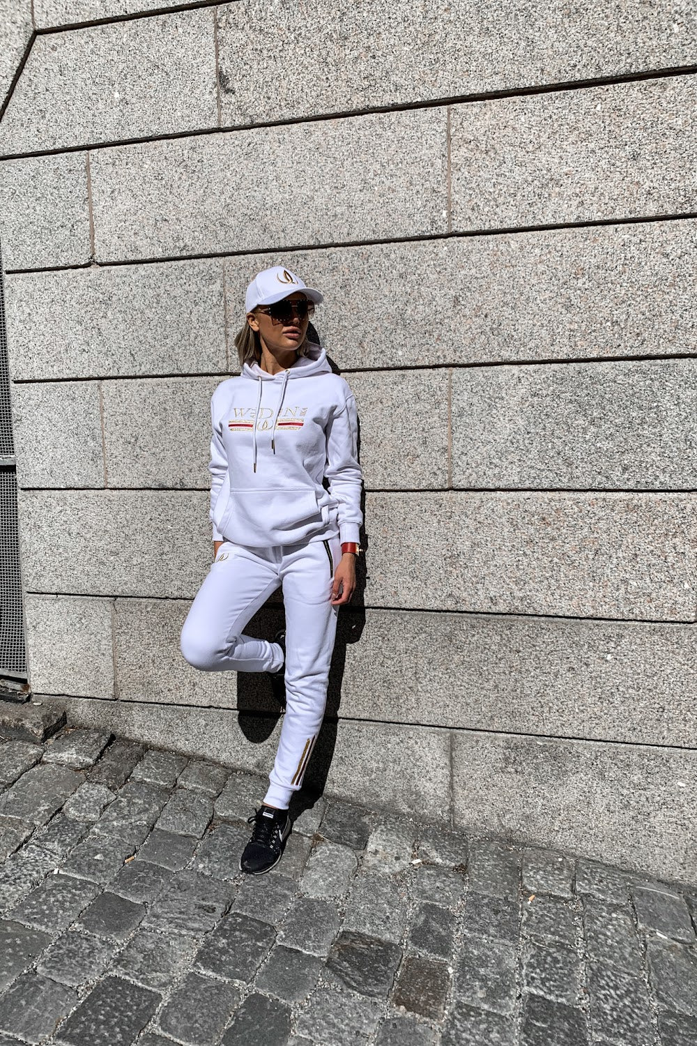 Tegen de stenen muur leunt het model met één voet opgetrokken, in een witte hoodie en witte joggers.