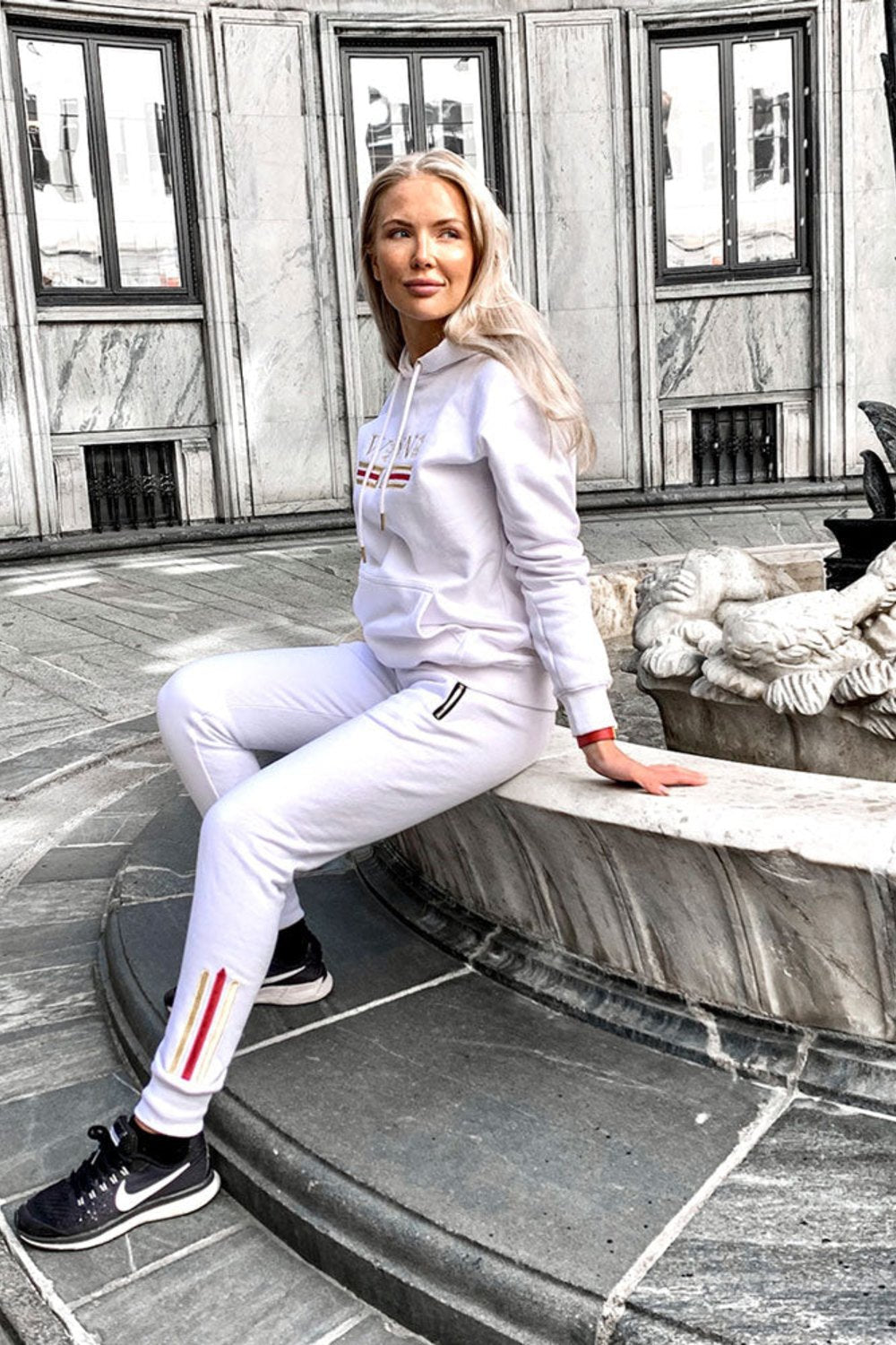 Het model zit bij een fontein in de stad in de witte hoodie en witte joggers met goudstreep.