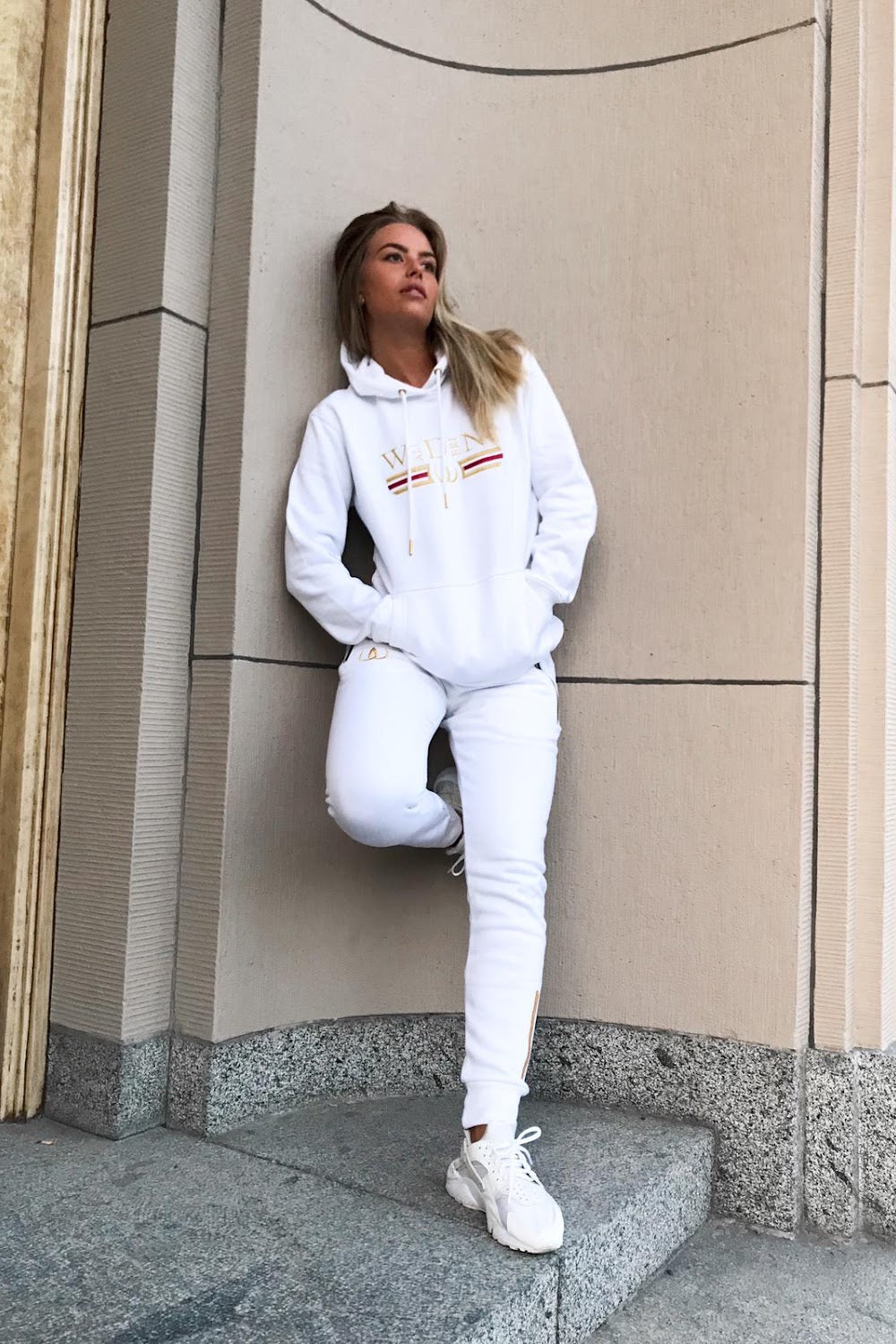 Tegen de huismuur staat het model in een witte hoodie en witte joggers, met witte sneakers.