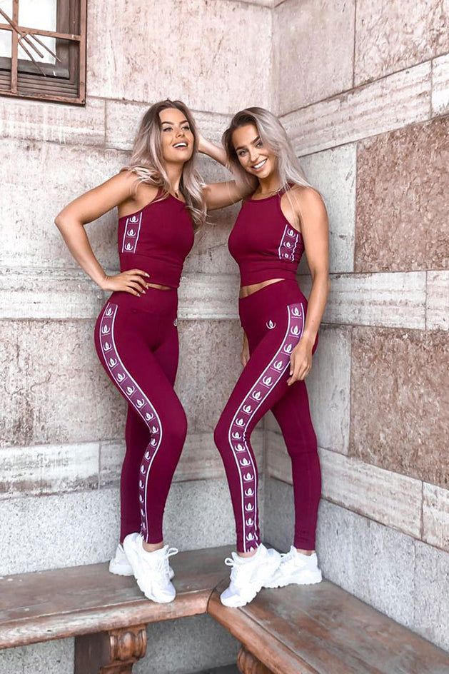 Twee modellen tegen een stenen muur geleund in wijnrode croptops en leggings met witte logobanden.