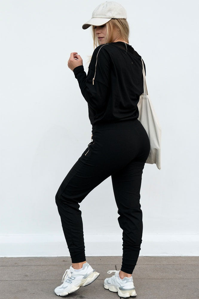 De broek met een zwarte sweater, pet en canvas tas. Bij de schouder is een goudkleurige rits te zien.