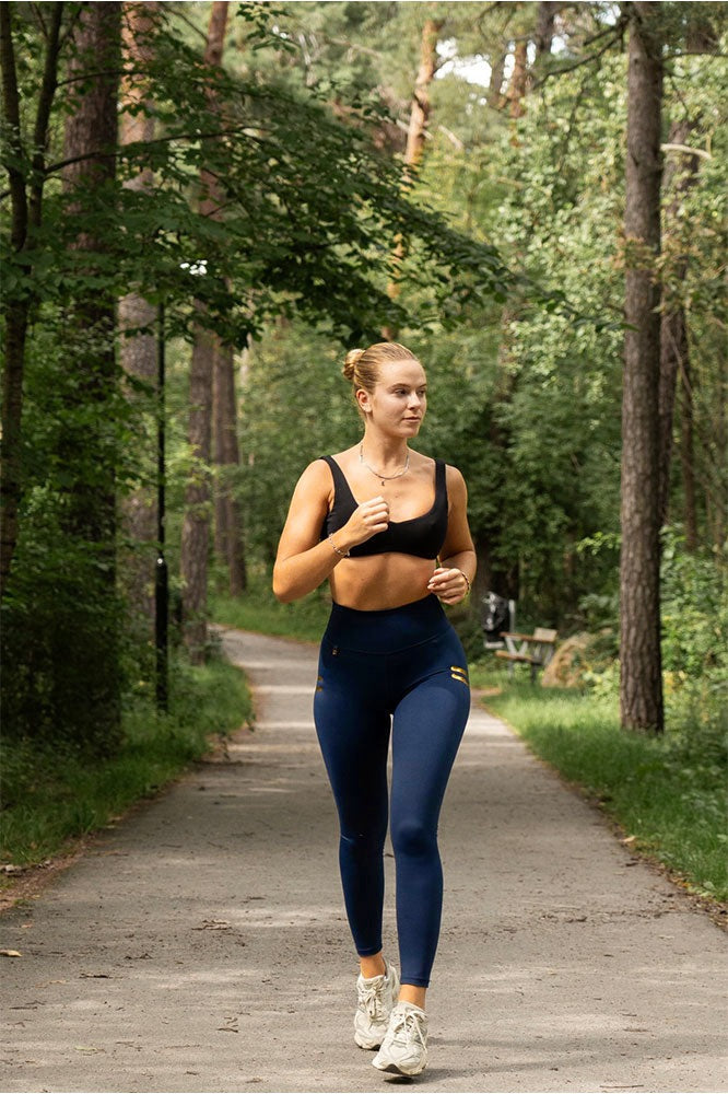 Hardlopen op een bospad in een marineblauwe legging met gouden strepen en een zwarte sportbh.