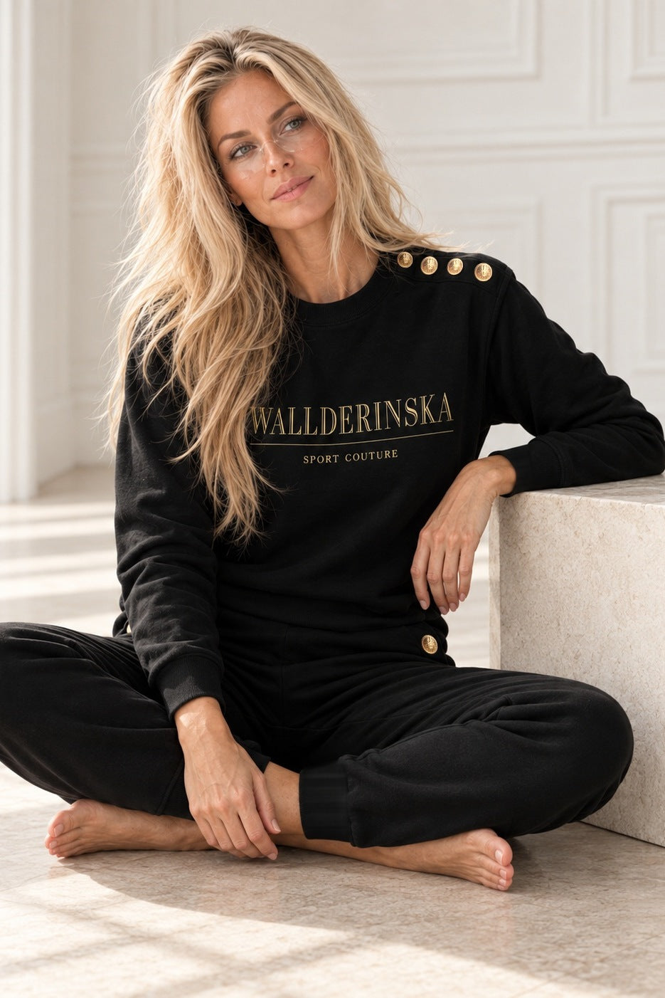 Edge Gold Button Sweater och Pants i svart, modellen sitter i skräddarställning vid en ljus stenkub i vit studio.